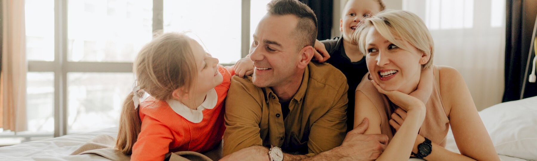 A happy family lies on a bed together: two kids and two adults smile and cuddle, enjoying a playful, cozy moment.