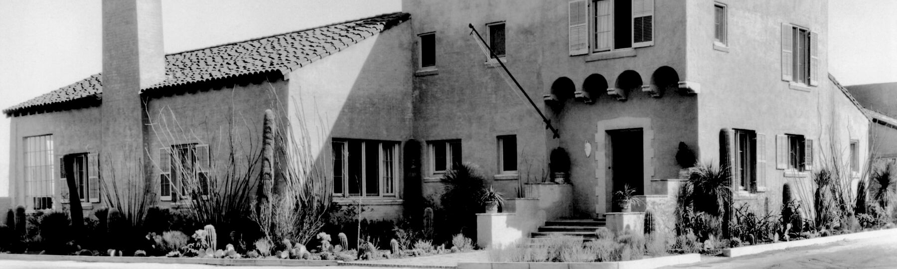 A black-and-white photo of a Southwestern-style house with stucco walls, arched windows, a tiled roof, courtyard plants, and a front street.