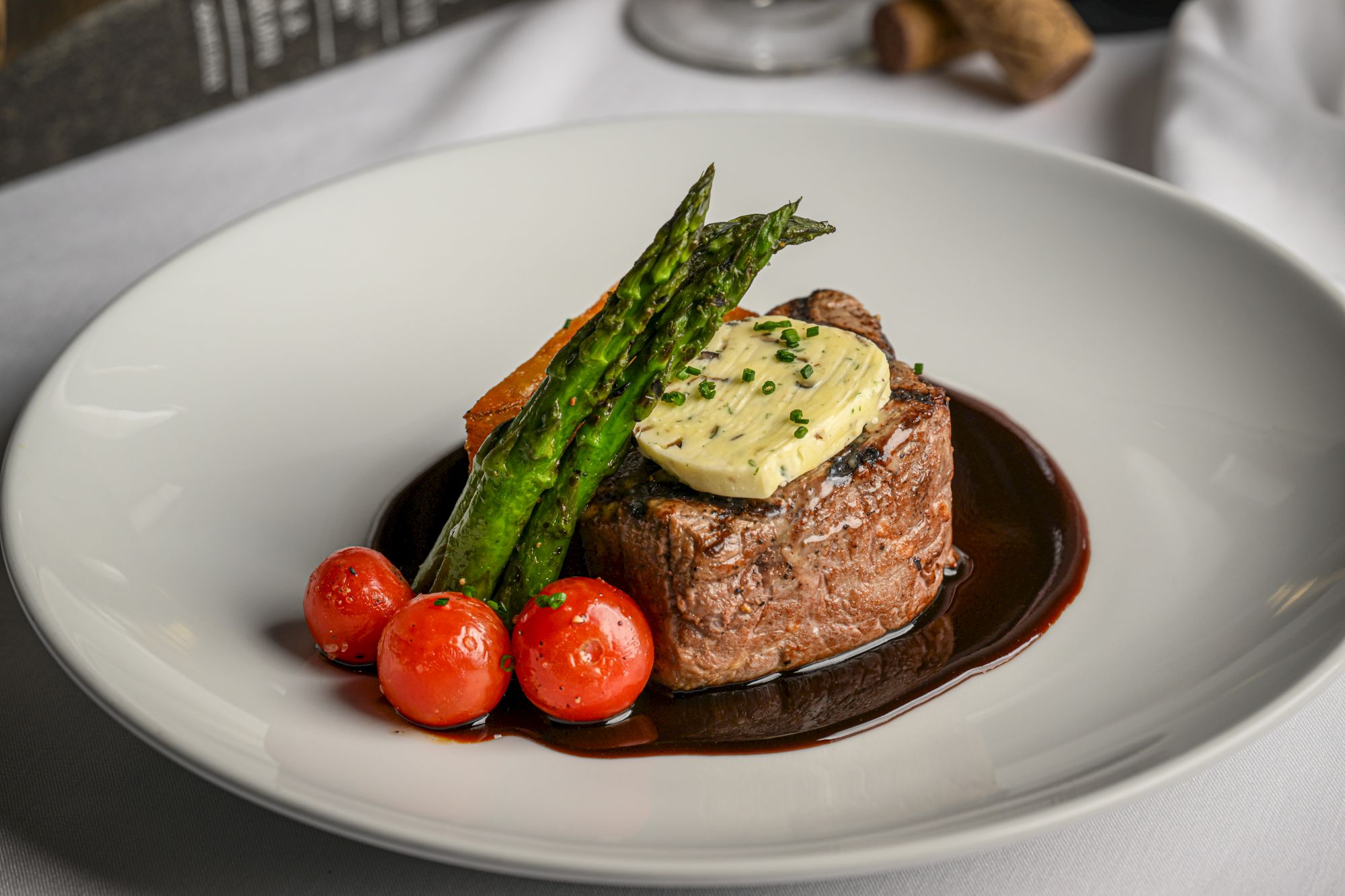 Prime rib steak on a plate with a dollop of herb butter, roasted asparagus, and cherry tomatoes, finished with a dark jus sauce.