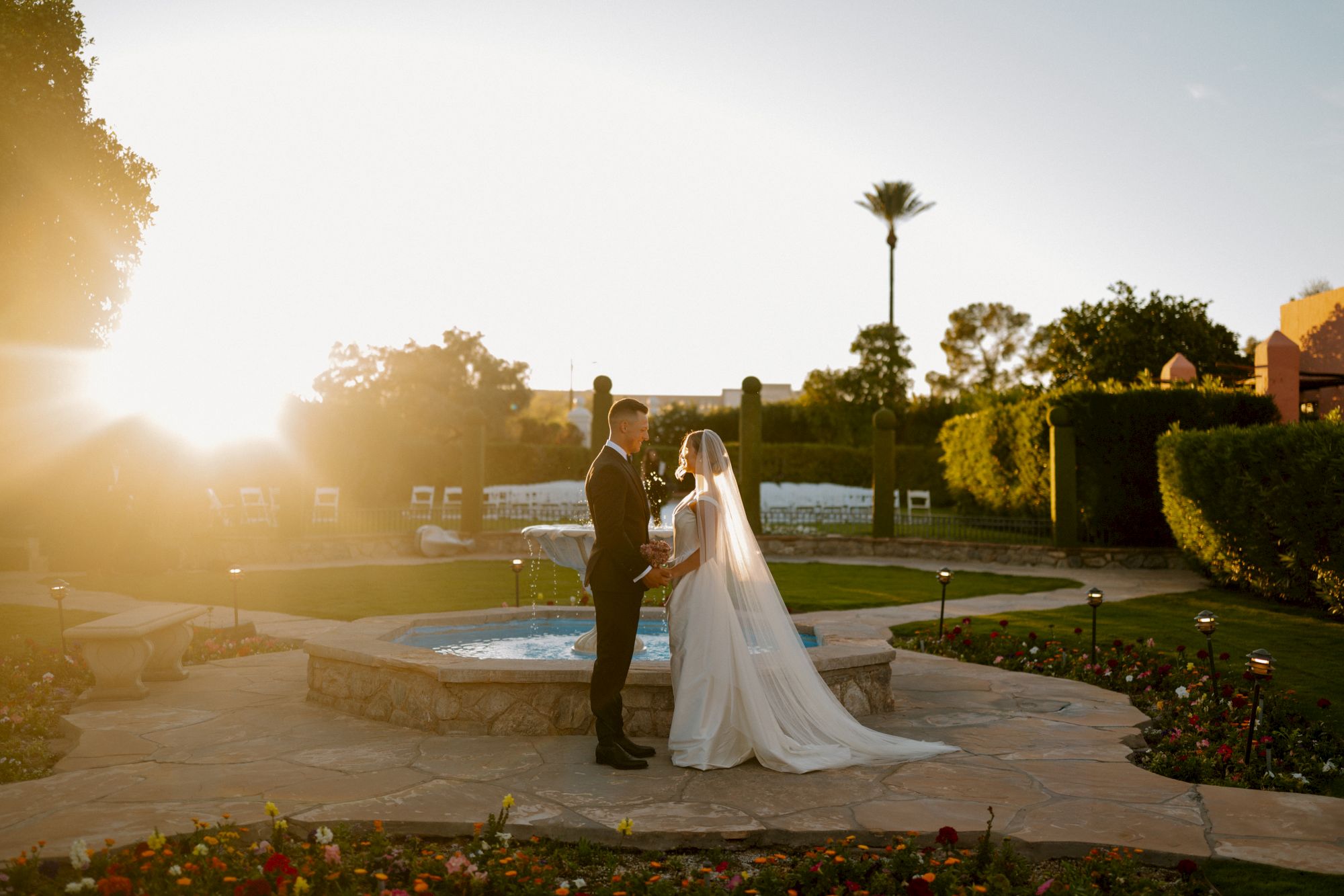 A bride and groom stand facing each other by a stone fountain in a sunlit garden, exchanging vows as soft light bathes the scene.