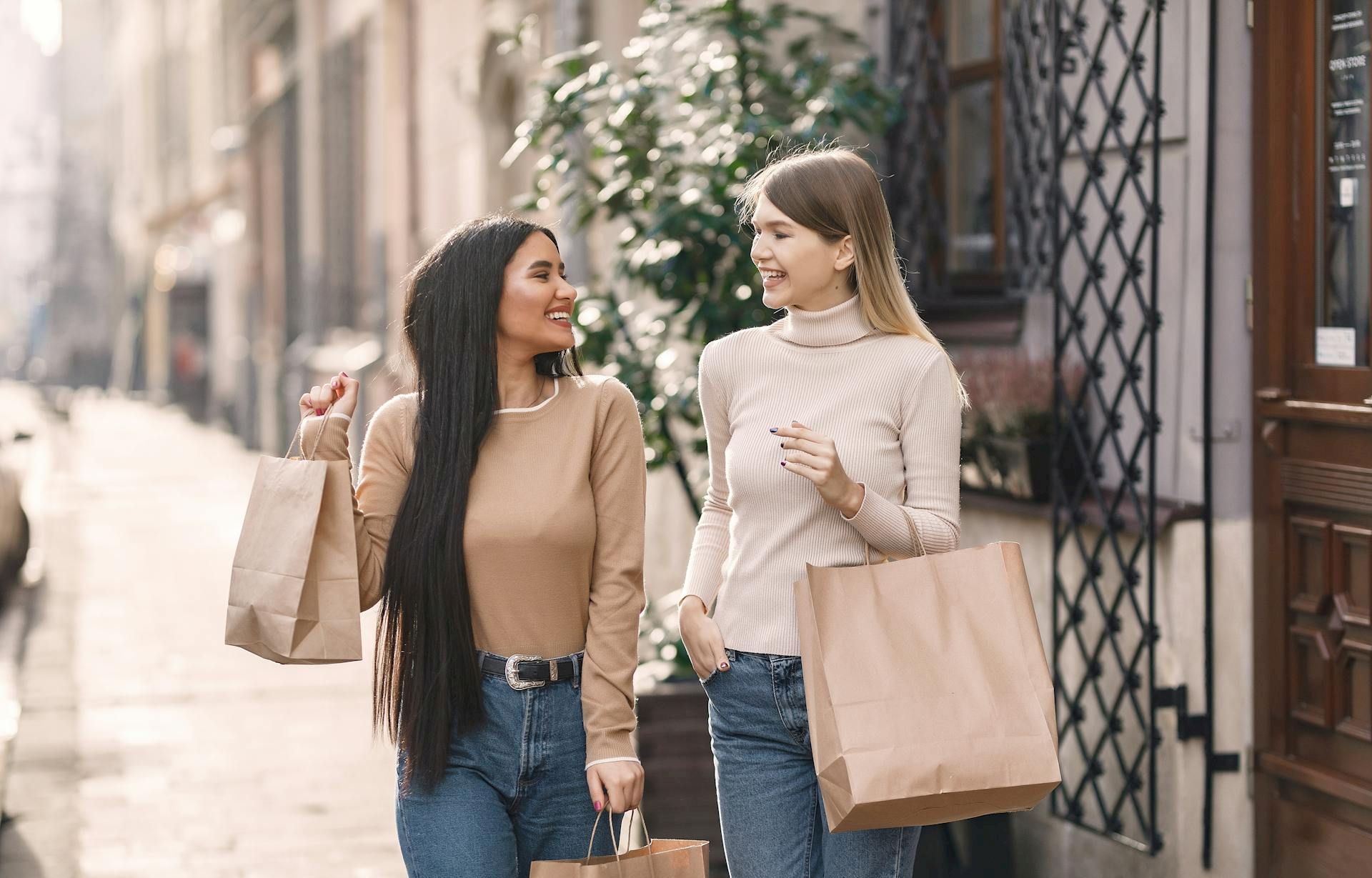Two women walking on a sunny street, shopping bags in hand, chatting and smiling as they stroll past shopfronts.