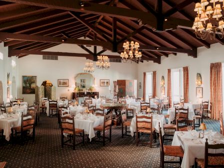 A cozy, elegant dining room with wooden beams, chandeliers, and white-tablecloths scattered across multiple round tables.