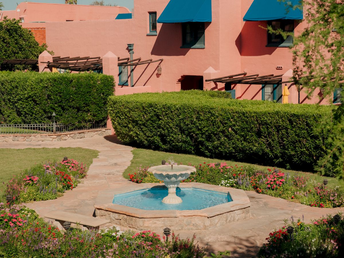 A pink coastal-style building with blue awnings, a small fountain, and lush greenery in a sunny courtyard.