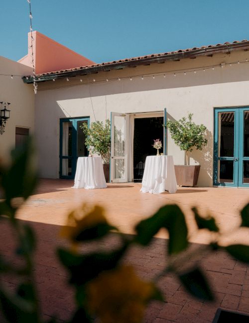 Two people stand behind small round tables on a sunny patio outside a pale villa with blue-framed doors, potted plants on each table.