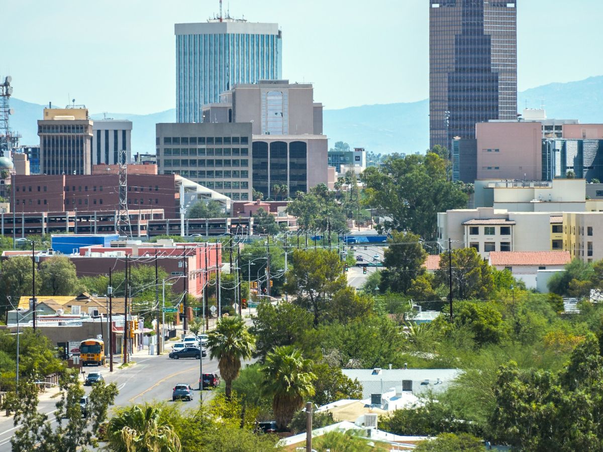 A city skyline with mid-rise buildings, trees in the foreground, and a clear blue sky, showing a mix of urban and green spaces, mostly daytime.