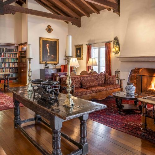 A cozy, traditional living room with a wood-beamed ceiling, fireplace, leather and upholstered seating, and a book-filled library nearby.