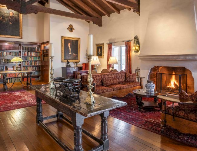 A cozy, traditional living room with a wood-beamed ceiling, fireplace, leather and upholstered seating, and a book-filled library nearby.