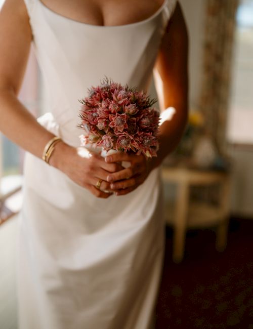 A bride in a white dress holds a small bouquet of dried pinkish-red flowers, standing indoors with soft light, bouquet close to waist. End with period.