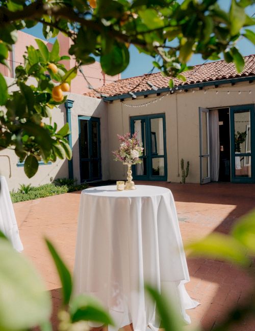 A small round table with a white tablecloth and a floral centerpiece stands in an outdoor courtyard, framed by greenery and a sunny villa.