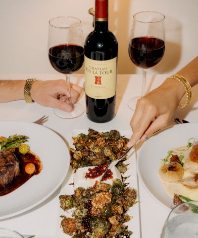 Two people share a table with red wine and a feast: a bottle center, three plates, and a skewered, crispy dish in the middle.
