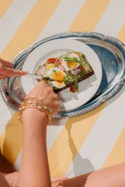 A person&rsquo;s hands place a small fancy toast topped with greens, eggs, and toppings onto a silver plate on a striped yellow background.