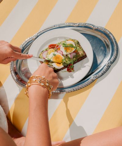A person&rsquo;s hands place a small fancy toast topped with greens, eggs, and toppings onto a silver plate on a striped yellow background.