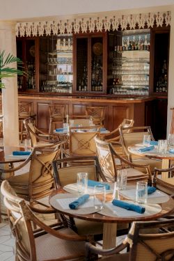 A cozy restaurant with wooden chairs, round tables, blue napkins, and a glass-front bar in the background, warm and inviting.