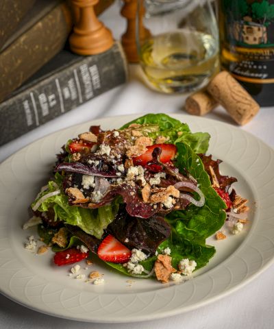 A fresh salad on a white plate: greens, strawberries, feta or goat cheese, nuts, and crumbled topping; wine and corked bottle in the background.