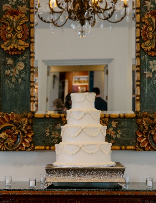 A tall white four-tier wedding cake sits on a decorative black-and-silver table, framed by an ornate mirror, with candles along the surface.