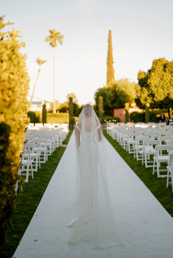A bride in a white wedding gown walks down a white aisle lined with chairs outdoors, flanked by trees, under a clear sky.