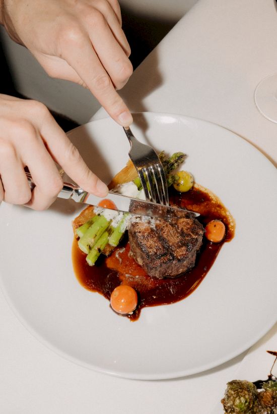 A person is cutting a plated steak with fork and knife, surrounded by a rich sauce, vegetables, and a wine glass on the table.
