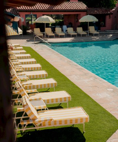 Rows of yellow-striped lounge chairs by a turquoise pool, with umbrellas and a pink building in the background. end.