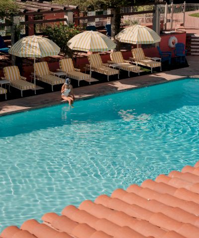 A sunny pool scene with clear blue water, sun loungers under striped umbrellas, and a person stepping into the pool, end it.