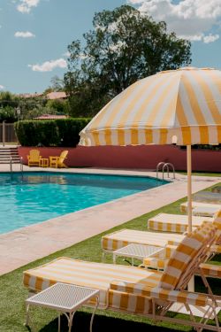 A sunny poolside scene with striped yellow and white lounge chairs, a matching umbrella, and a bright blue pool under a clear sky.
