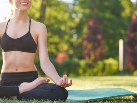 A person practicing yoga outdoors on a mat, meditating with legs crossed, wearing a black sports bra and leggings, sunny park setting.